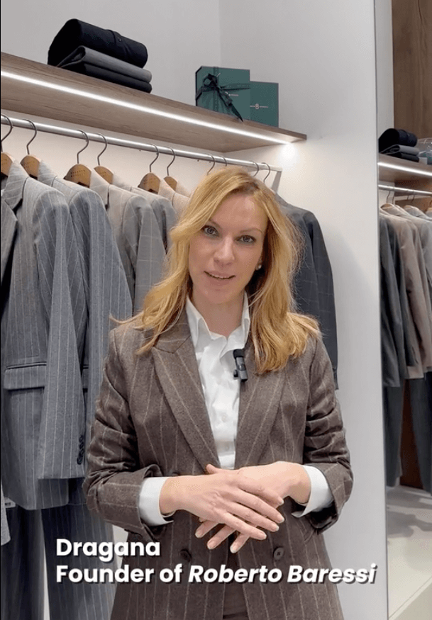 Dragana, standing facing a camera in a suit, with her hands crossed. Behind her is a row of suits on a hanger
