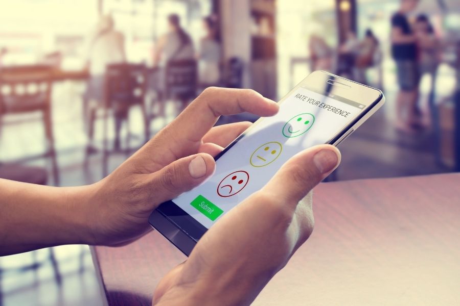 Two hands holding a phone. Phone displays 3 smiley faces, good, neutral and sad. And says rate your experience.